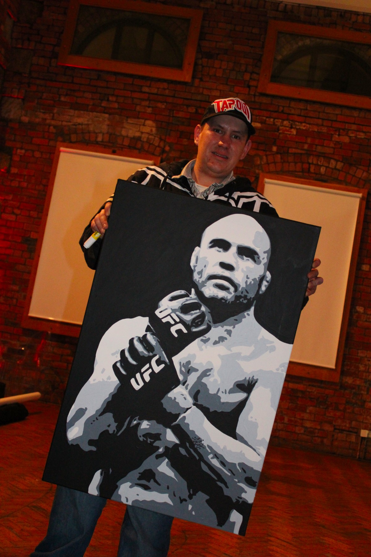 An artist in a Tapout cap holding a large black and white pop-art canvas painting of Randy Couture in Leeds