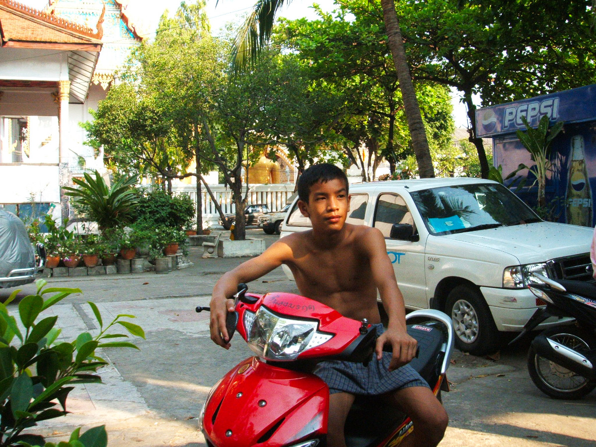 A young Thai Nak Muay boy sitting on a red motorbike outside the camp, temple and golden Buddha visible behind him