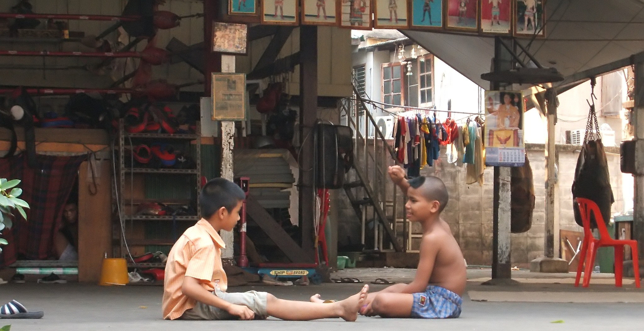 Two young Nak Muay Thai boys sitting on the gym floor at Saengmorakot Camp, training equipment around them