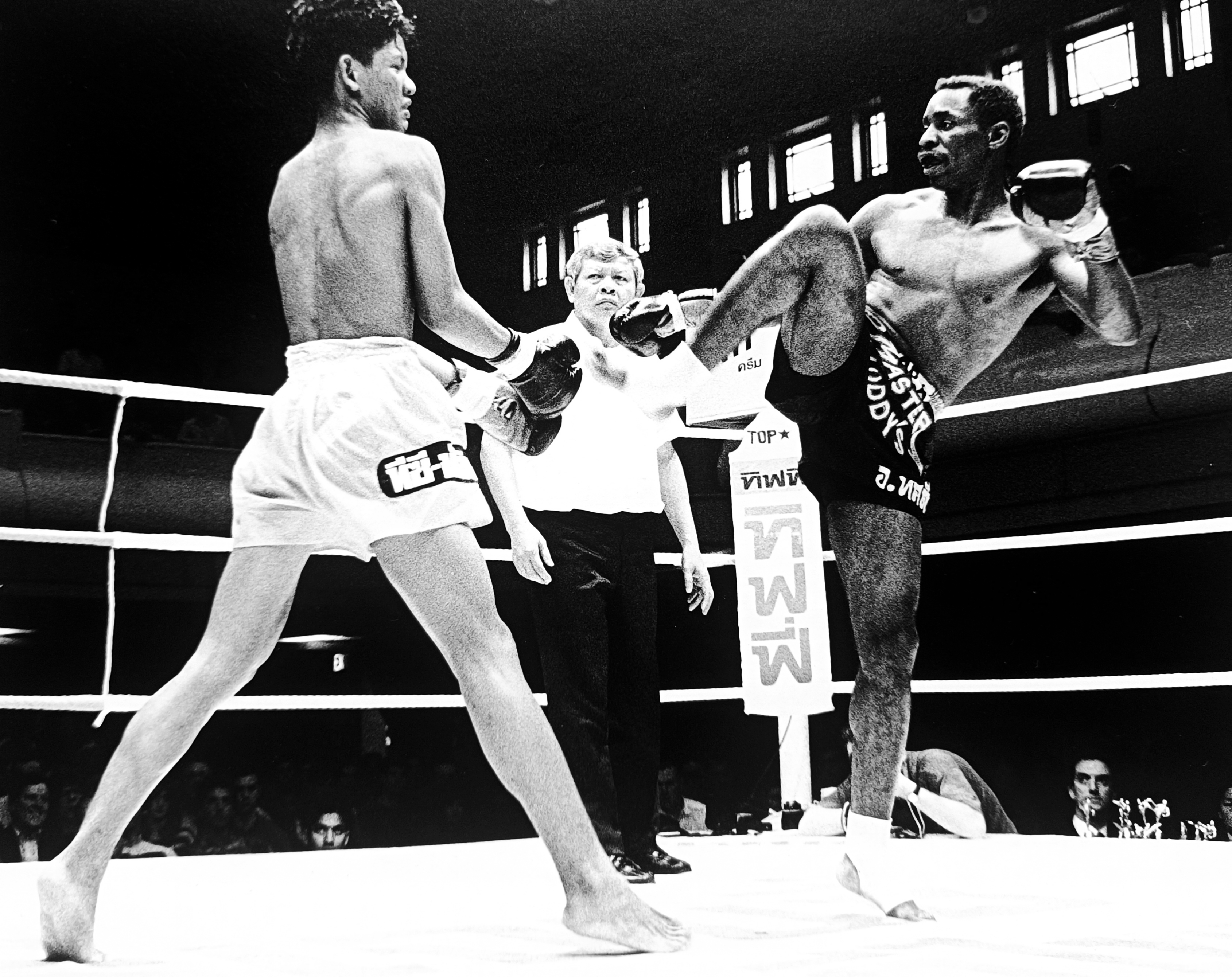 Ronnie Green in Master Toddy's shorts throwing a devastating high kick at his Thai opponent, referee watching, Thai advertising banners visible