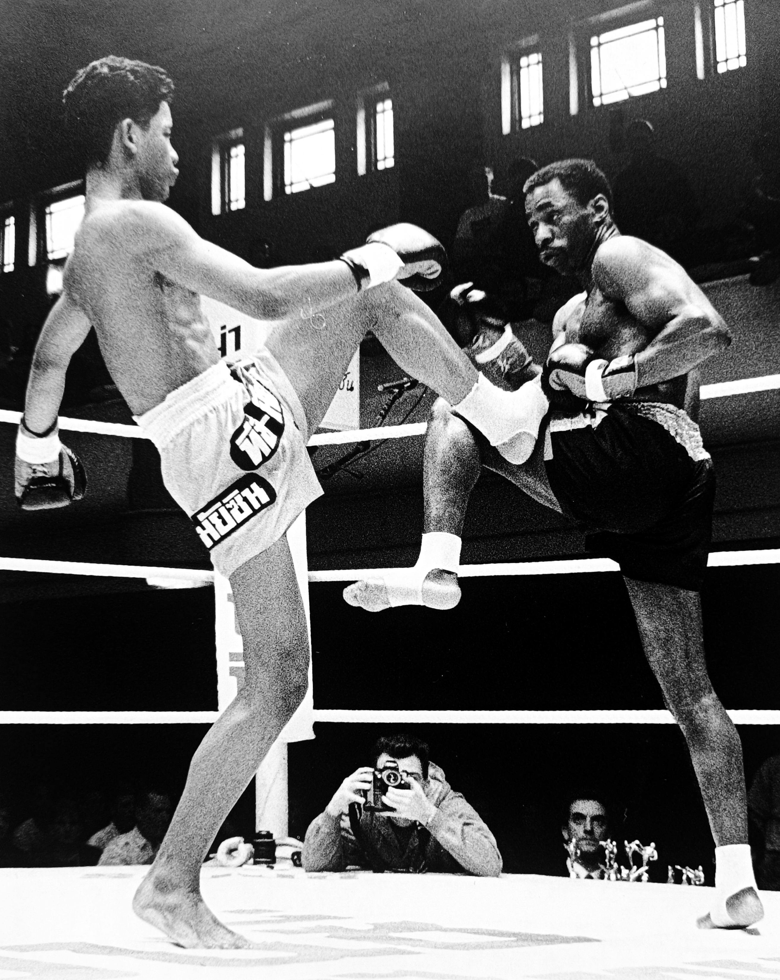 Ronnie Green blocking a kick from his Thai opponent, press photographer visible ringside capturing the action