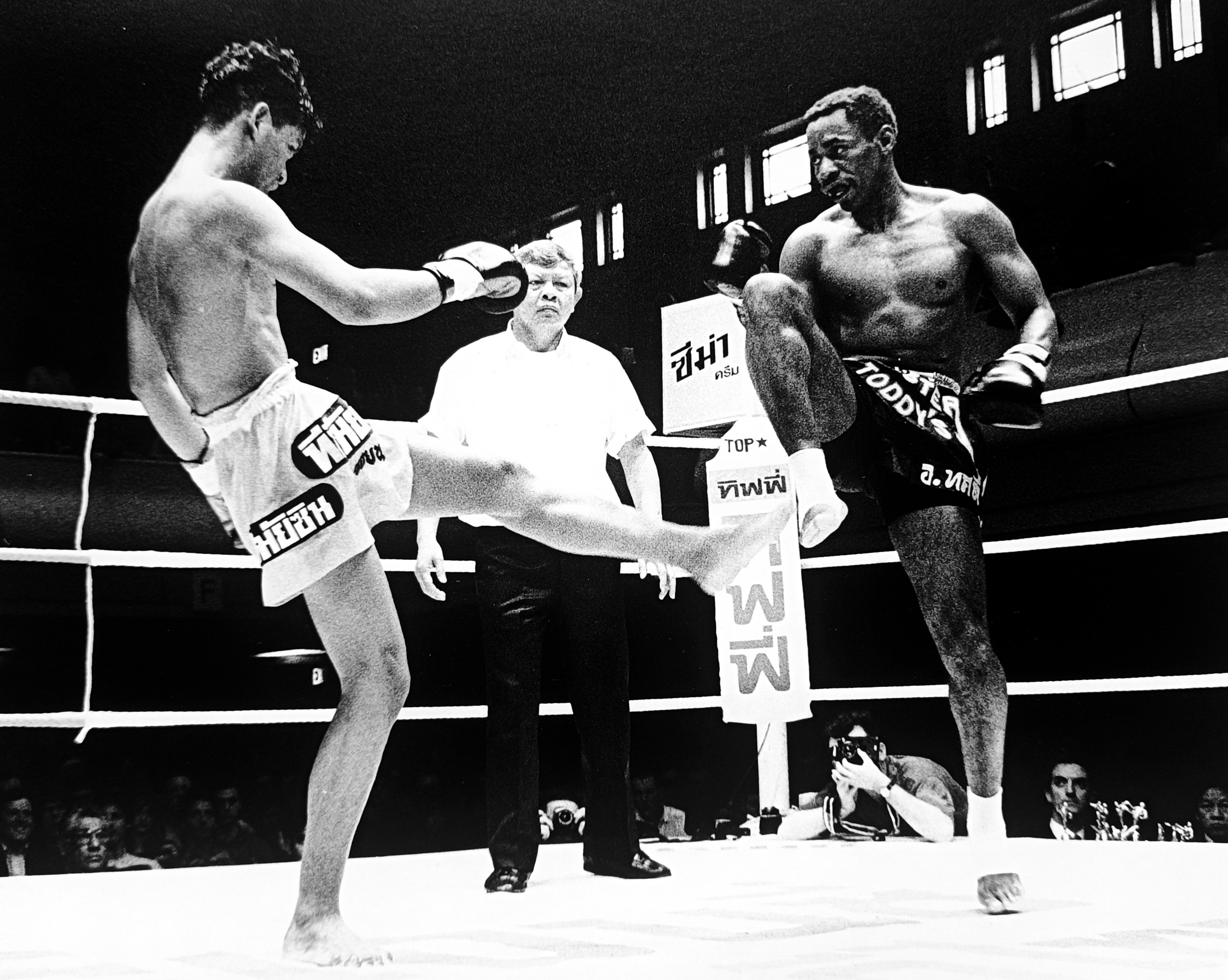 Ronnie Green and his Thai opponent exchanging kicks, referee between them, photographer visible ringside