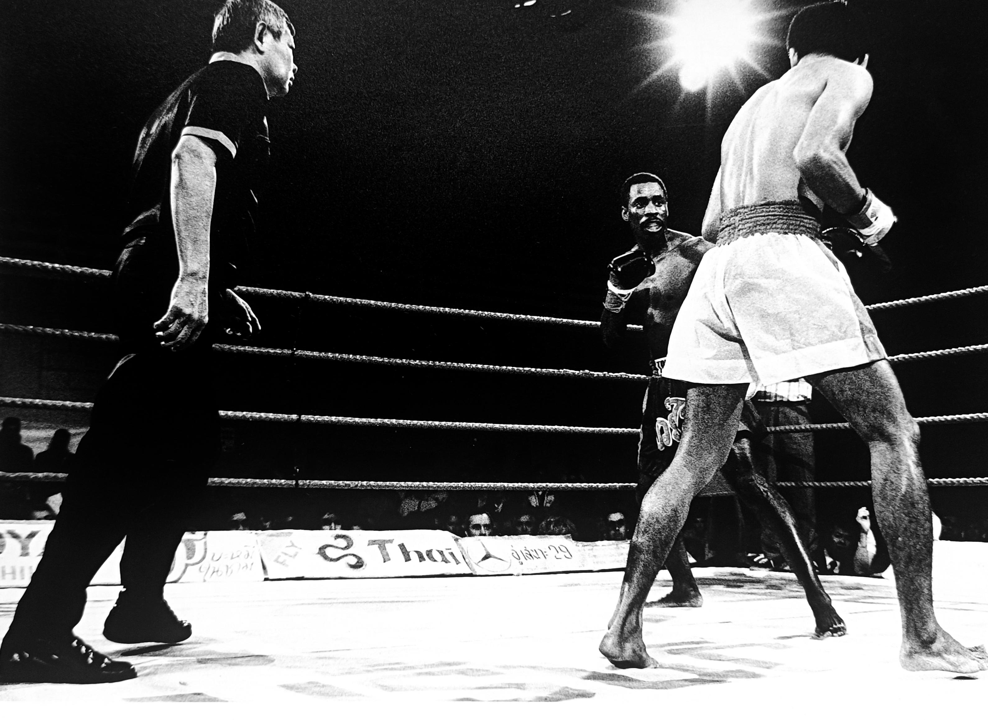 Ronnie Green facing his Thai opponent under a single spotlight, referee stepping in, low angle dramatic shot