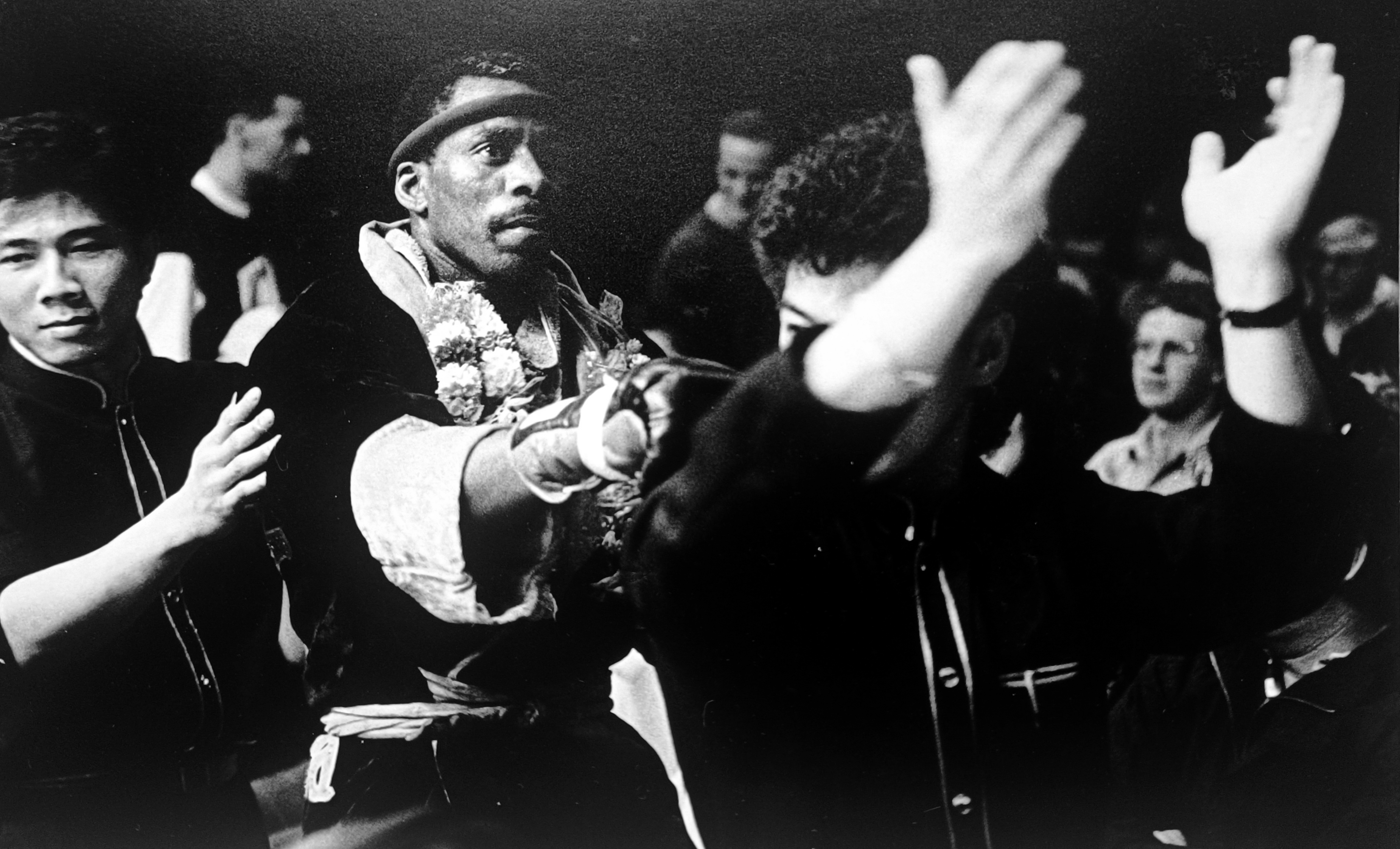 Black and white photograph of Ronnie Green victorious after a fight, flowers around his neck, arm raised, crowd behind him