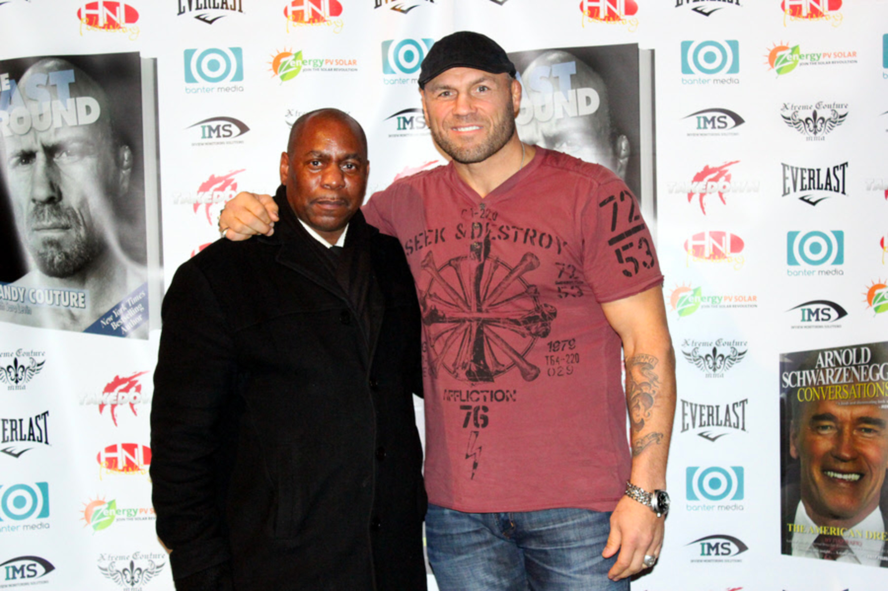 Ronnie Green and Randy Couture standing together in Manchester, Ronnie's home town, with The Last Round poster and Xtreme Couture branding behind them