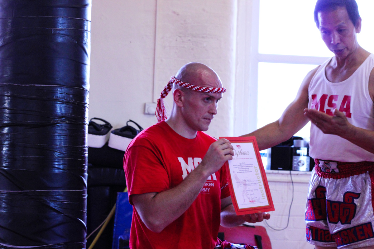 Grandmaster Sken presenting Raphael with his Muay Thai instructor diploma, Raphael wearing a Mongkhon headband and red MSA shirt