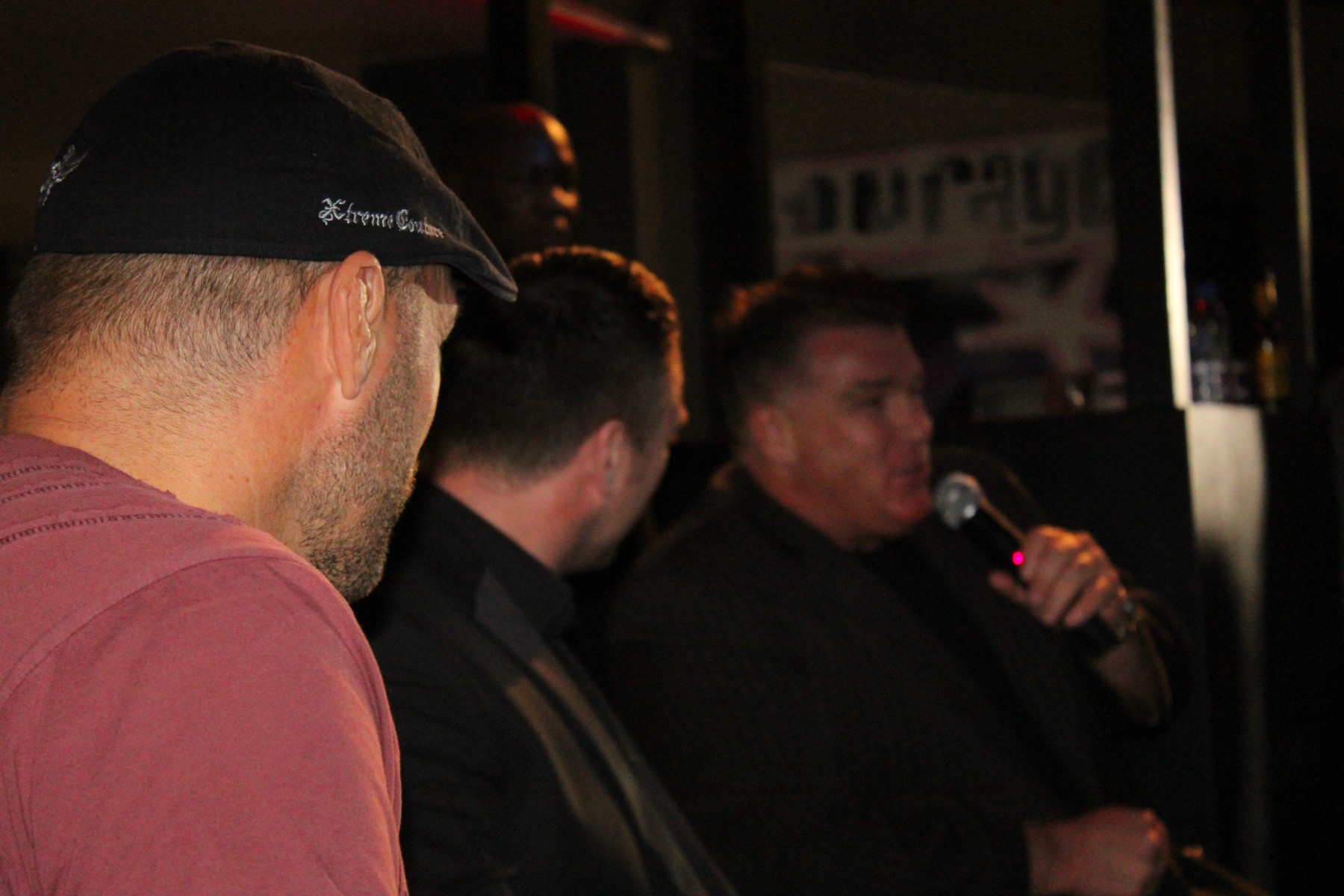 Three world-class champions in one frame: Randy Couture foreground, MC in middle, Big Joe Egan on mic, and Master Ronnie Green hidden in bokeh as bouncer